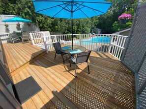 A wooden deck with a table and chairs, a blue umbrella, and a pool in the background.