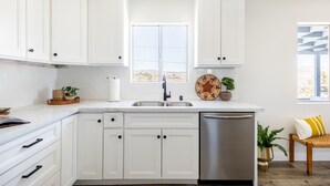 A modern kitchen with white cabinets, a stainless steel sink, and a dishwasher. There is a window with a view of the outdoors. - Boho🎨 Haus✦NearPark✦Views✦HotTub✦🎮Den✦🔥Pit✦Ev🔋 (Joshua Tree)