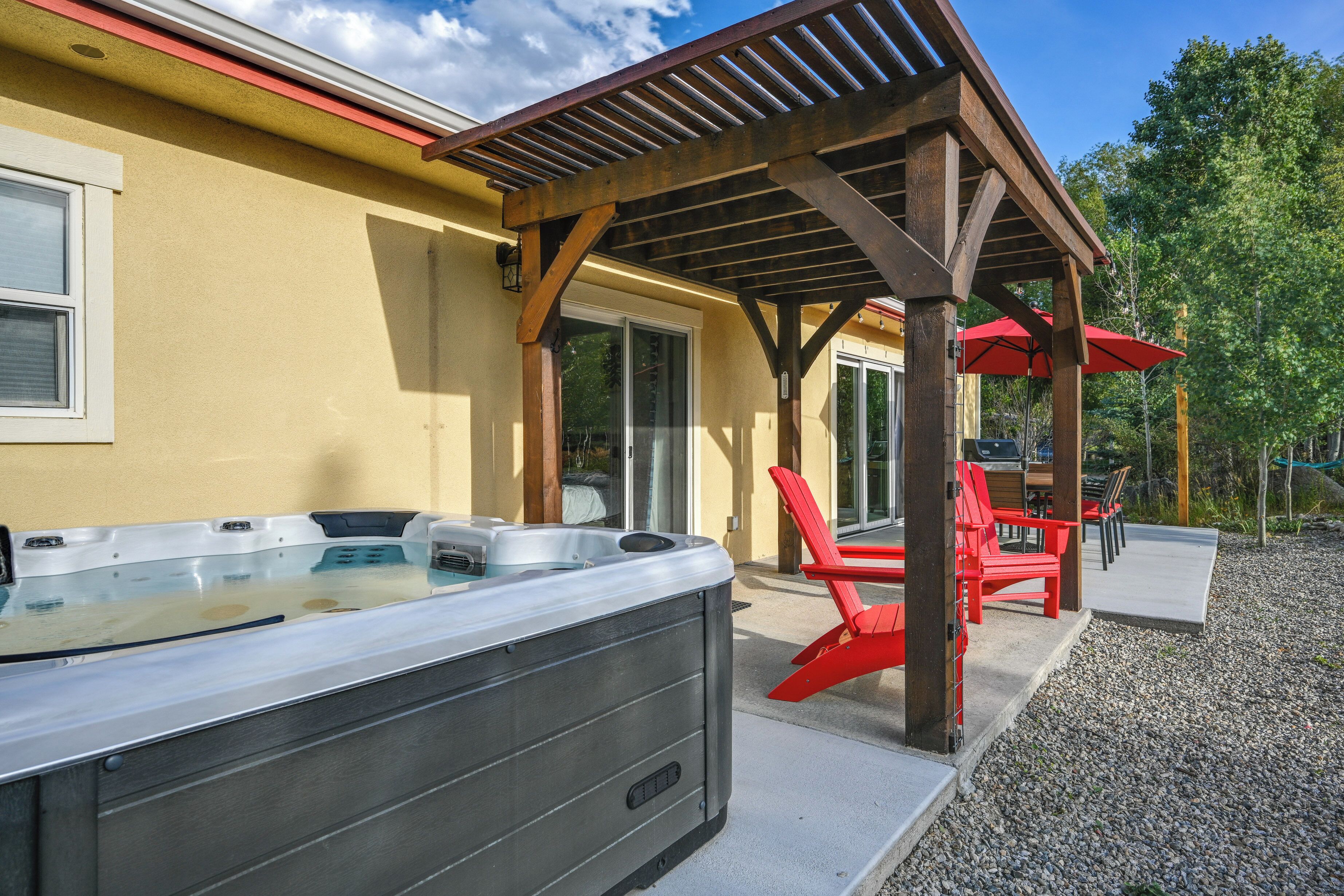 Een hottub onder een houten pergola met rode stoelen en parasols, een terras met grind en een huis met een gele gevel.