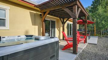 Een hottub onder een houten pergola met rode stoelen en parasols, een terras met grind en een huis met een gele gevel.