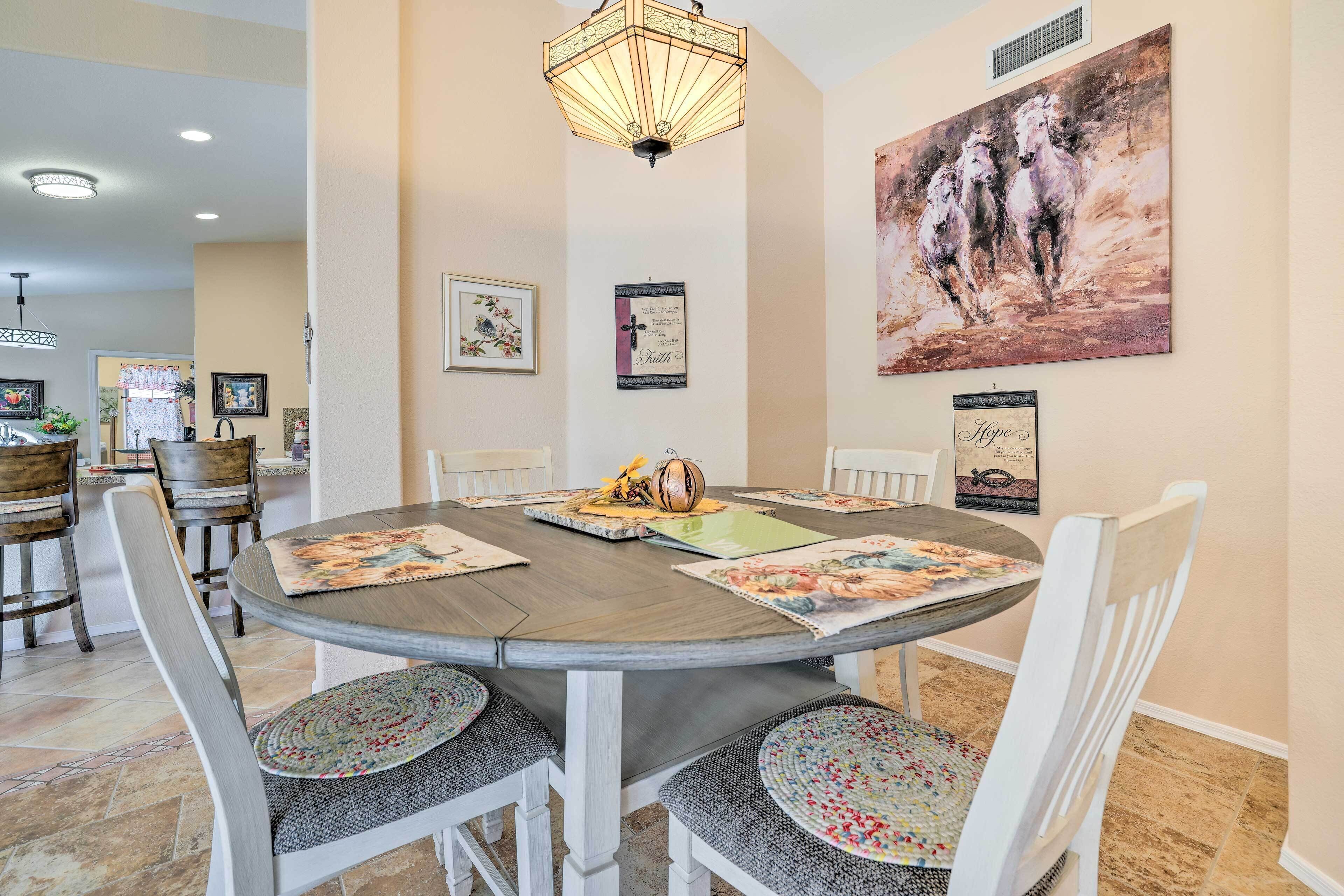 A dining area with a round table, white chairs, and a chandelier. There are framed pictures on the walls and a painting of horses.