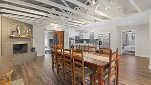 A spacious dining area with a long wooden table and chairs, a brick fireplace, and a kitchen in the background. - Serene Getaway Near Baylor and Magnolia Perfect for Relaxation and Exploration (Waco)