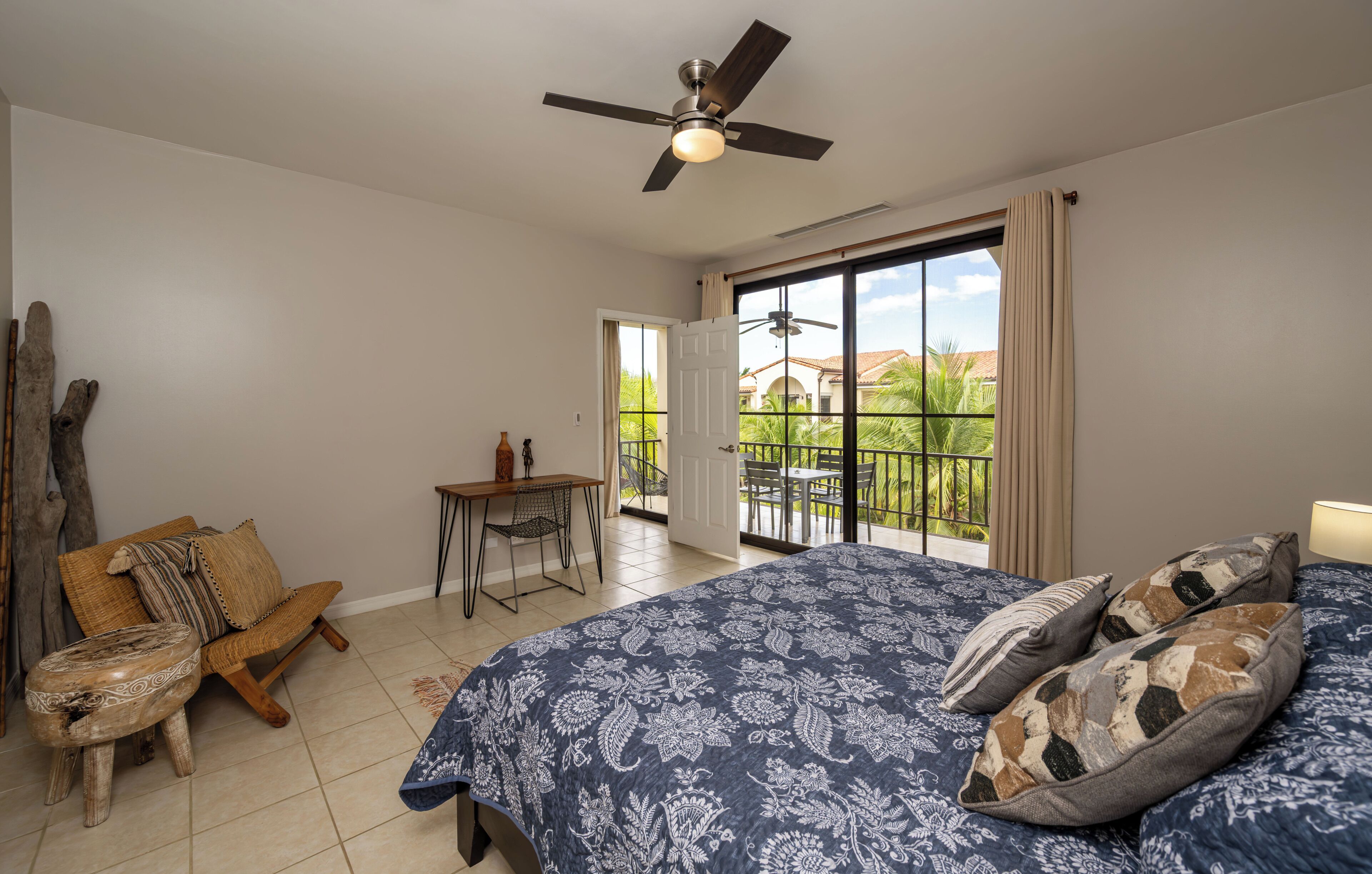 A bedroom with a bed, a ceiling fan, a small table, a chair, and a view of the outside.