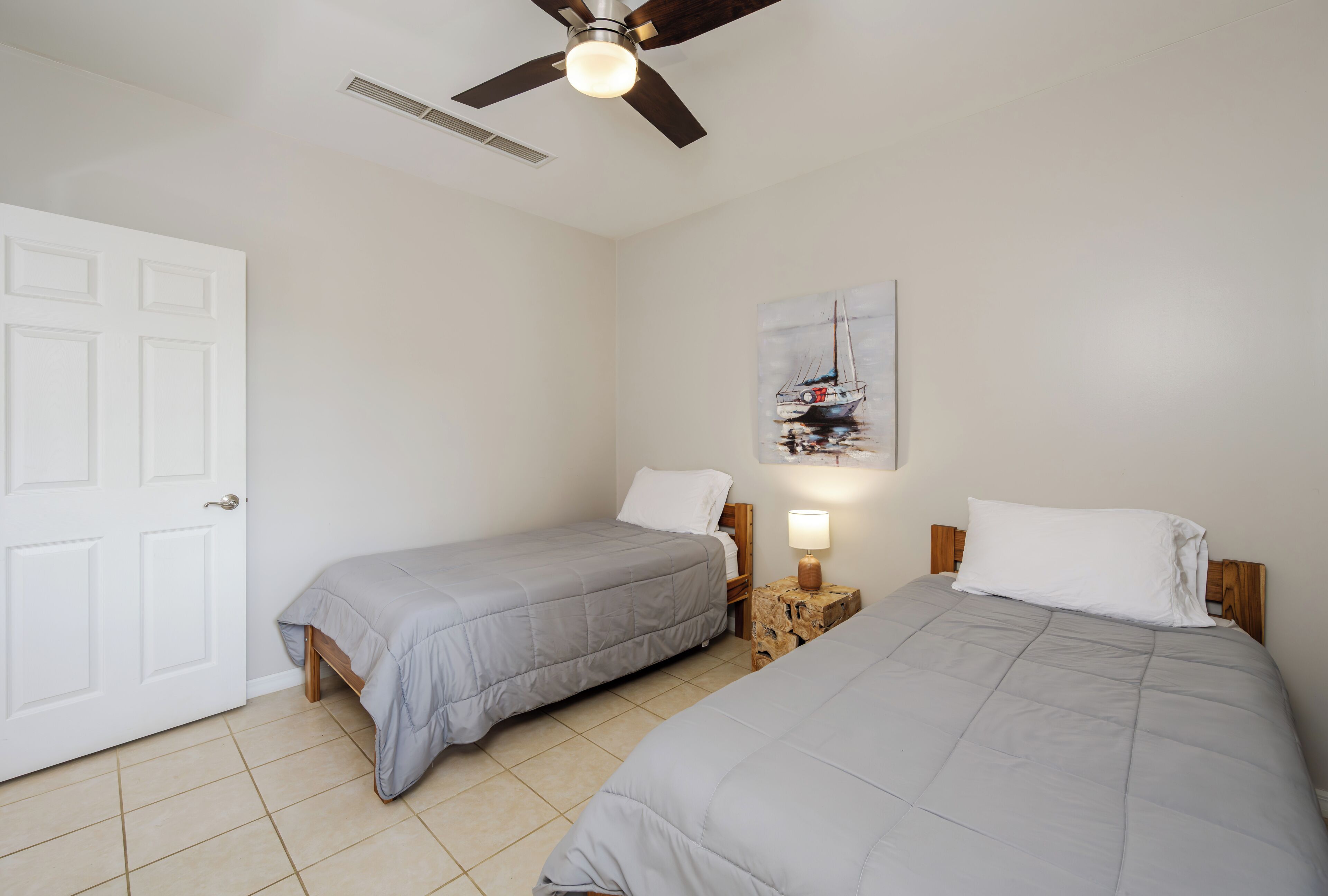 A bedroom with two beds, a ceiling fan, a painting, and a door.