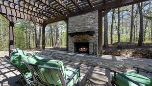 Un patio exterior con chimenea de piedra, sillas acolchadas verdes y una estructura de pérgola.