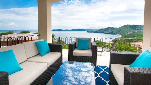A balcony with wicker furniture, a glass table, and a view of a coastal landscape.