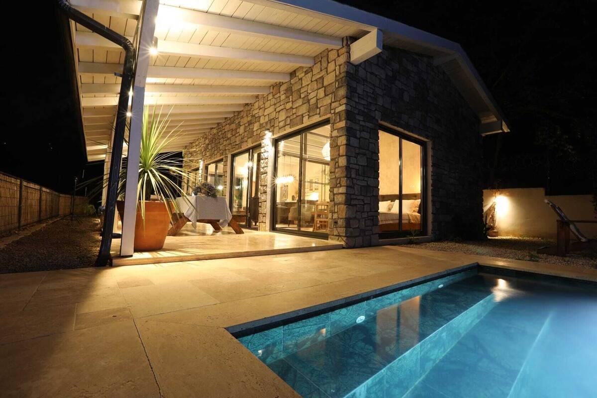Ein modernes Haus mit Naturstein-Fassade, Glastür, Swimmingpool und einer Terrassenfläche mit Tisch und Stühlen.