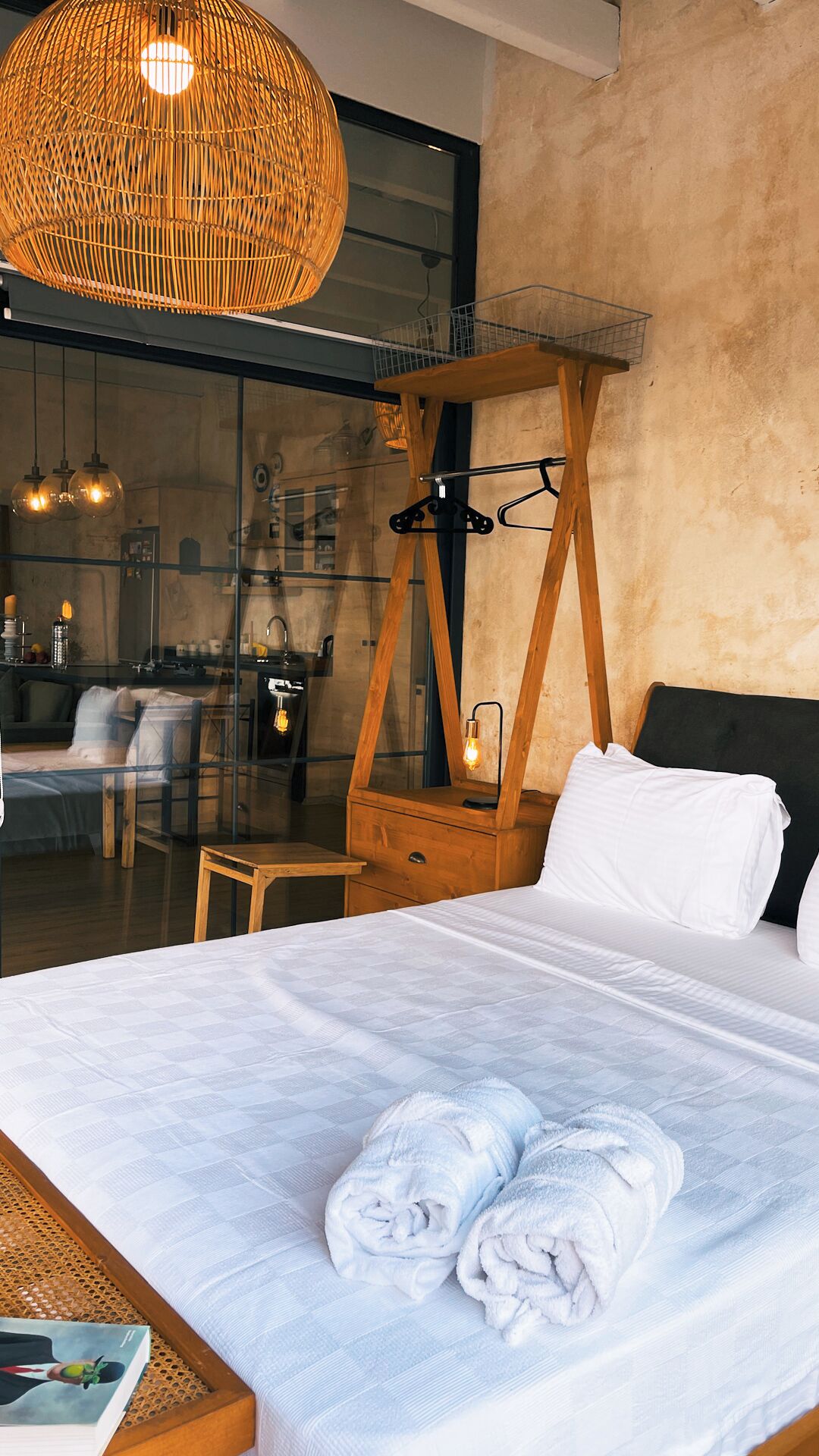 Una habitación de hotel moderna con una cama grande, cabecera de madera, lámpara colgante y vistas del interior del hotel a través de un muro de cristal.