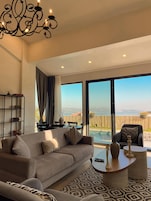 A spacious living room with a large sofa, a coffee table, and a bookshelf. A large window offers a view of a pool and the ocean.