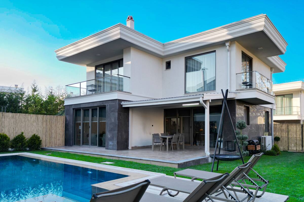 Ein modernes, zweistöckiges Haus mit Swimmingpool, Sitzgelegenheiten im Freien und einer überdachten Terrasse.