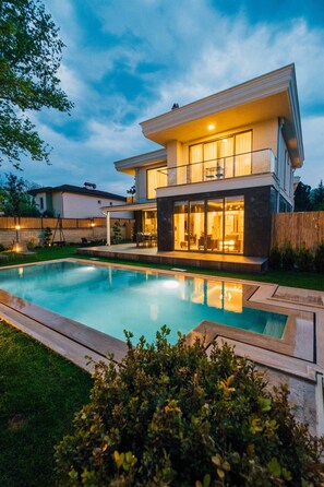 Ein modernes, zweistöckiges Haus mit Swimmingpool, überdachter Terrasse und gepflegtem Rasen.