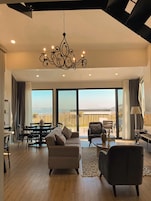 A spacious living room with a large window, a chandelier, and a view of the beach.