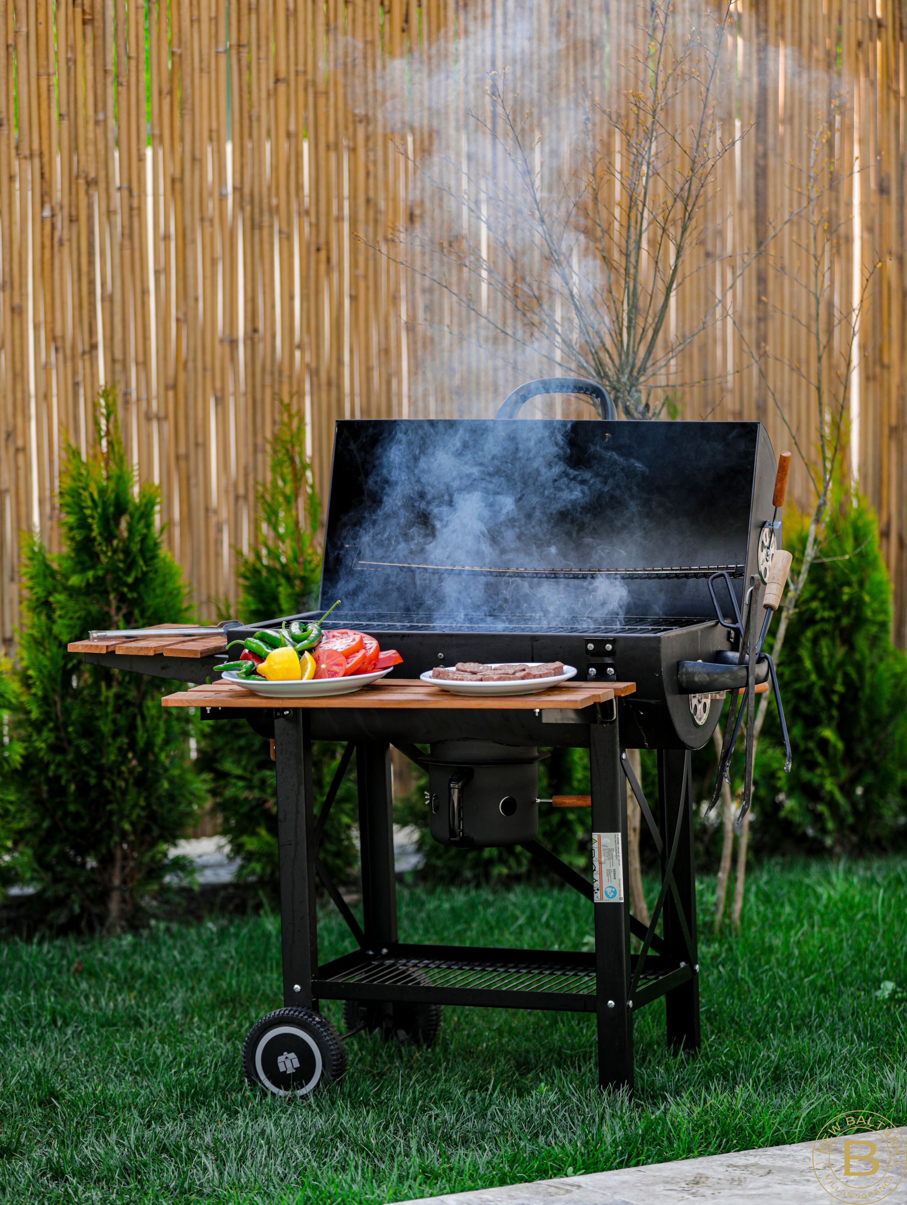 Una parrilla portátil con asa, un plato de verduras y una olla sobre un soporte con ruedas en un área cubierta de césped, con una cerca de bambú al fondo.