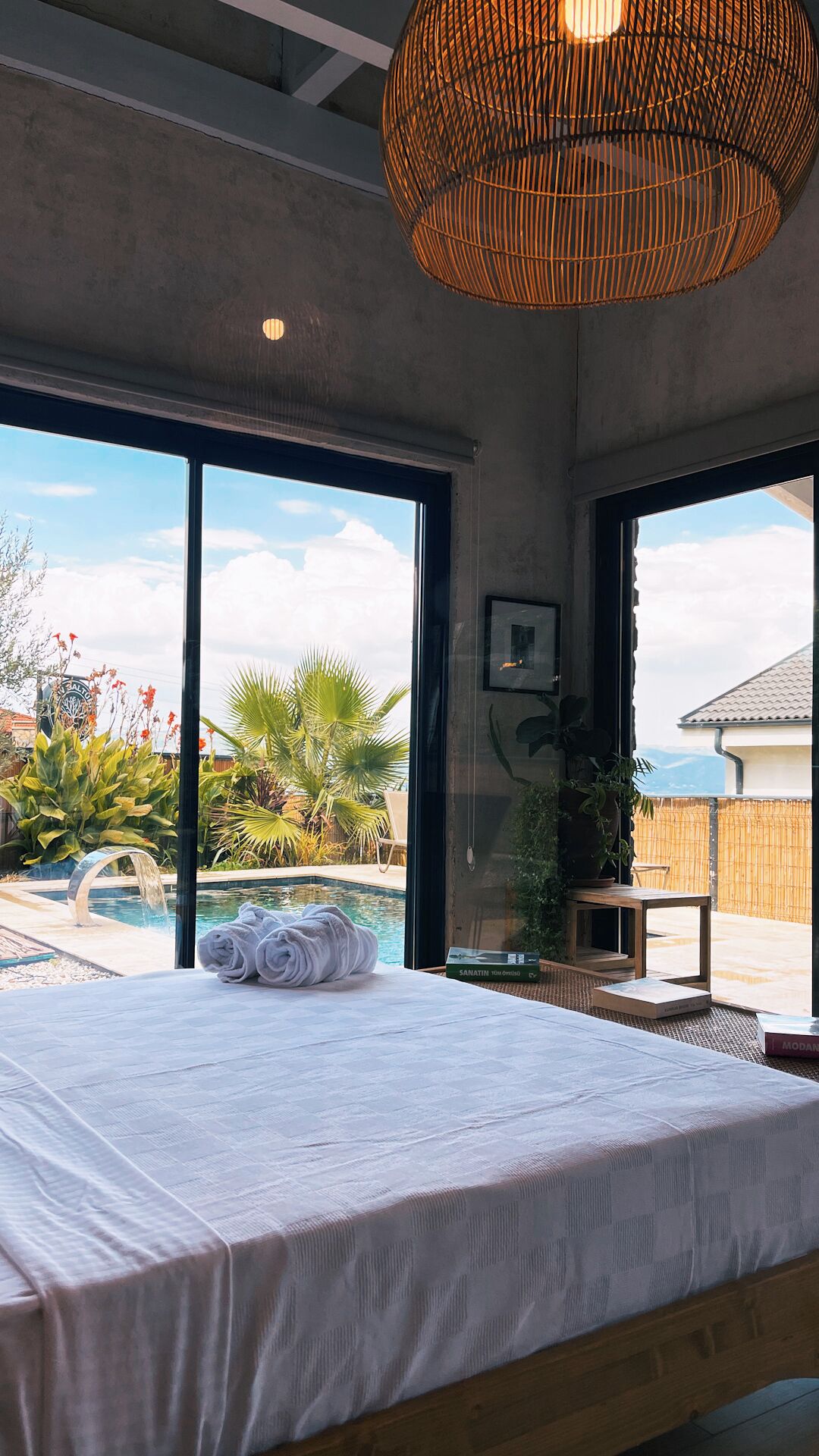 A bedroom with a large bed, a view of a swimming pool, and a wicker ceiling light.