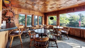 Un'accogliente ambientazione rustica in legno, con tavoli e sedie rotonde, ampie finestre che si affacciano su un lago e alberi, e un'area bar con un bancone in legno e una lampada.