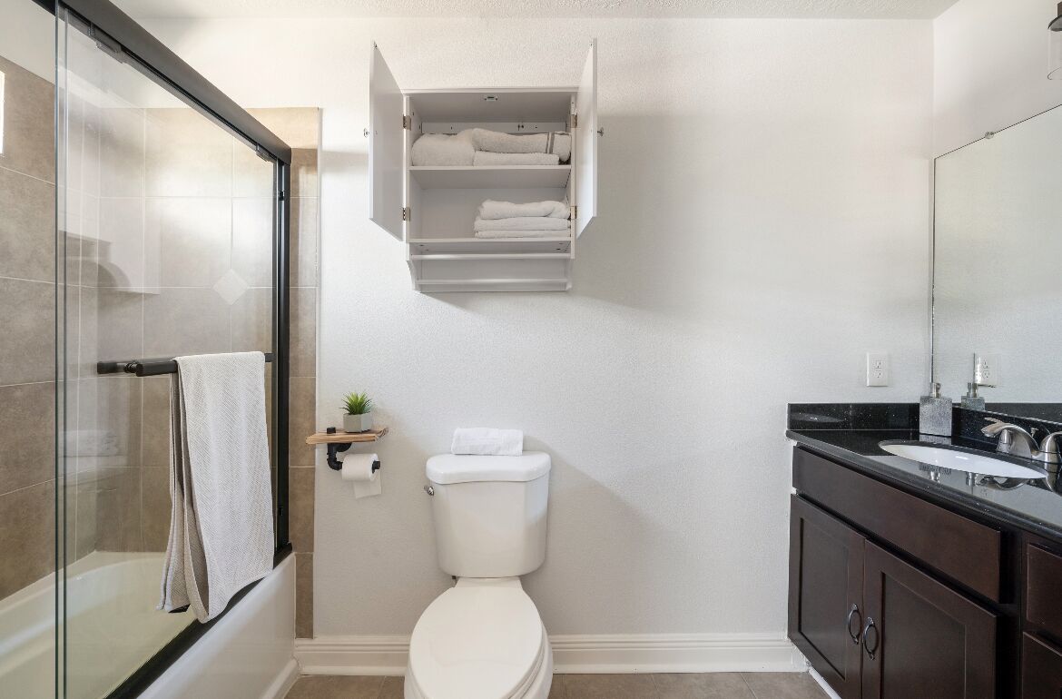A bathroom with a glass shower enclosure, a toilet, a sink with a dark countertop, and a white cabinet above the toilet.