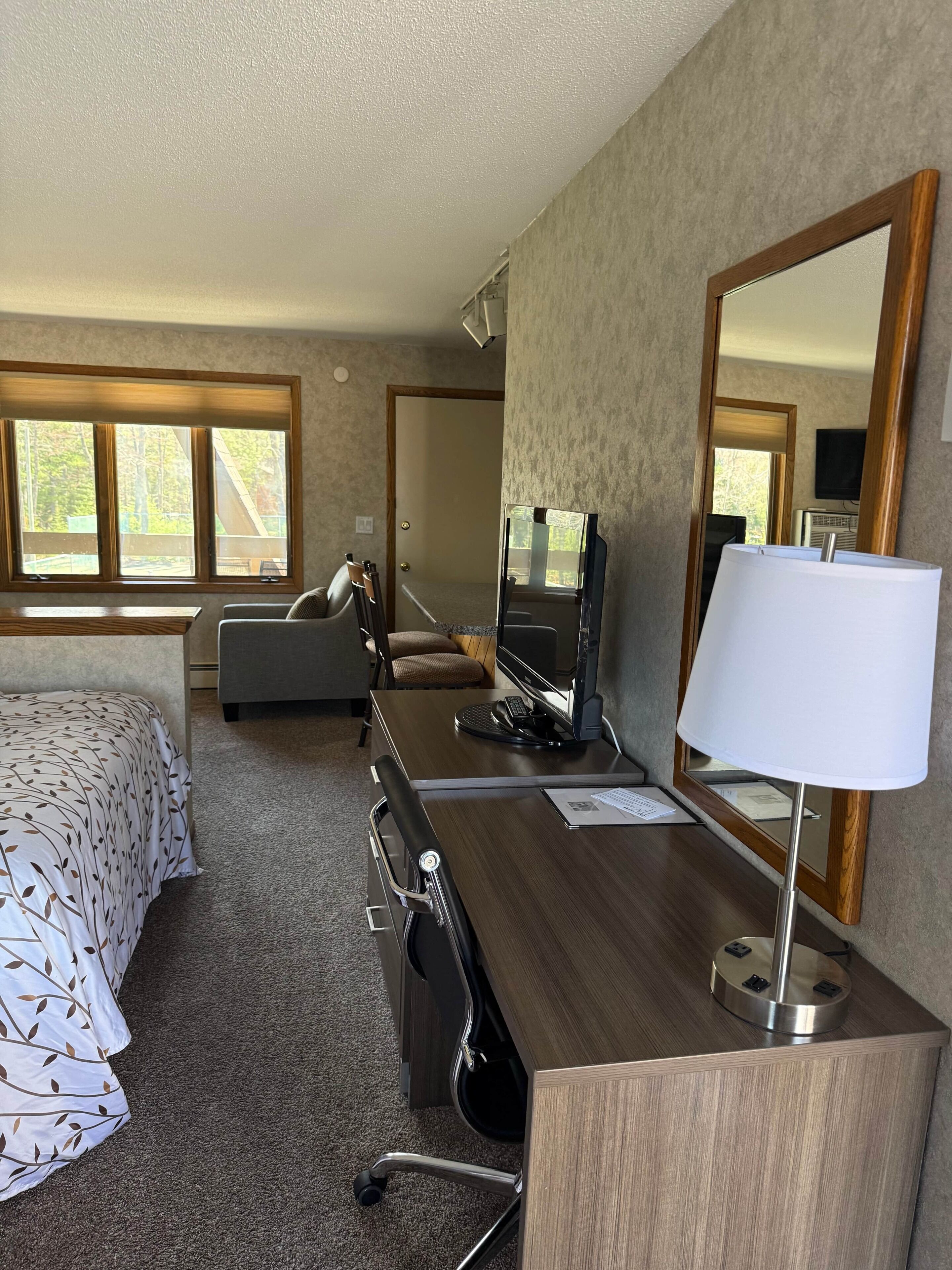 Quarto de hotel com cama, escrivaninha, computador, cadeira, luminária e um espelho grande.