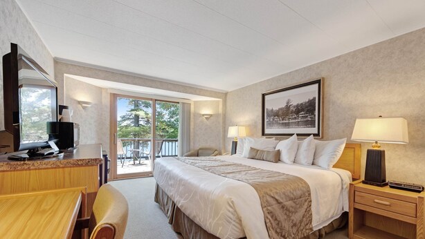 Habitación de hotel con una cama grande, un escritorio con una computadora, una silla, una mesita de noche con una lámpara y una ventana con vista a árboles.