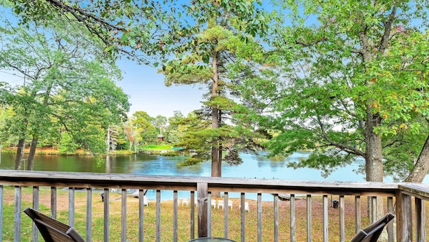 A wooden deck with a glass table and chairs overlooking a lake and trees.