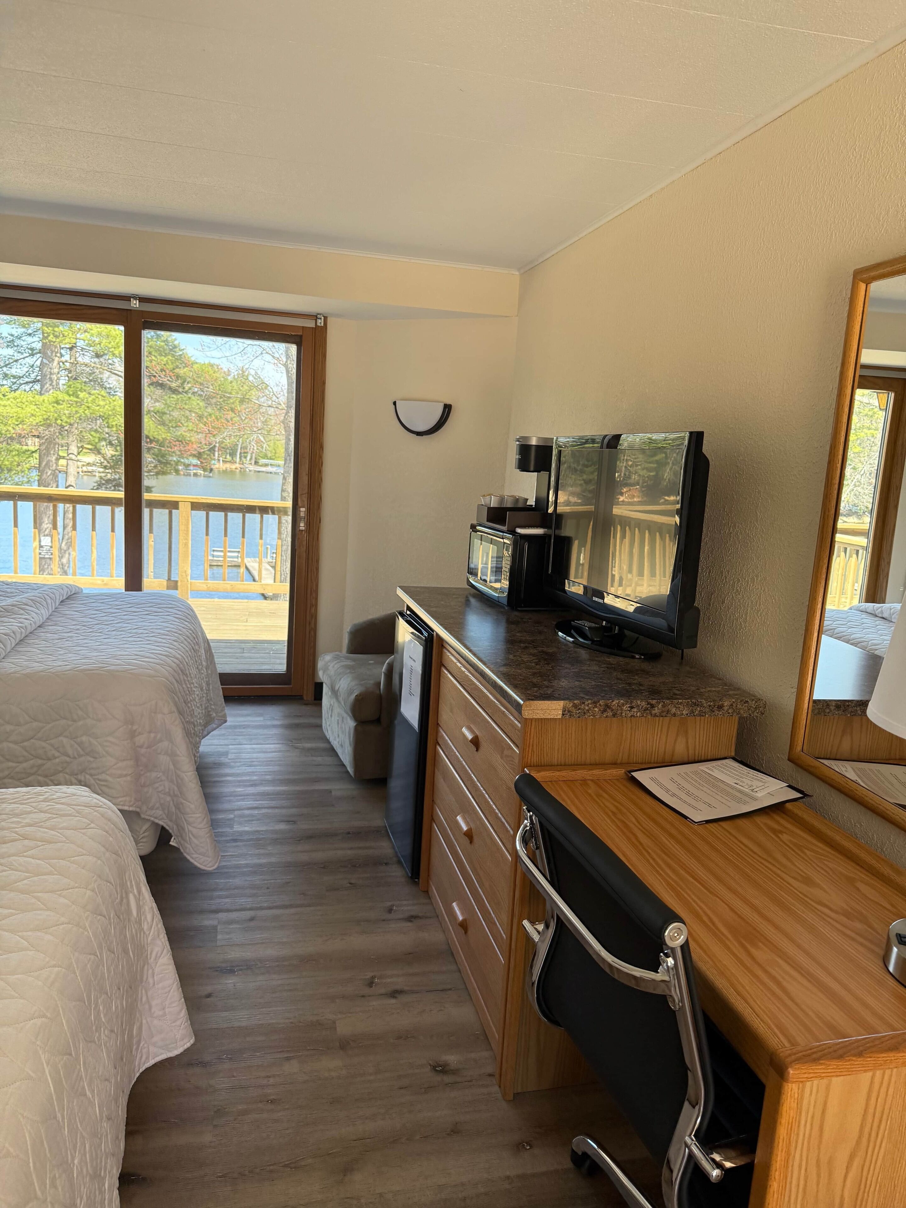 Ein Hotelzimmer mit zwei Betten, einem Holztisch mit Stuhl, einem Fernseher und Blick auf einen See durch große Fenster.