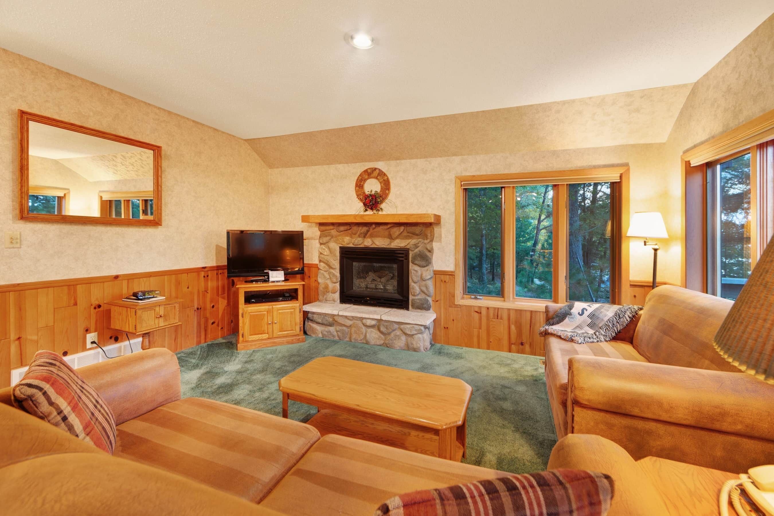 A cozy living room with a stone fireplace, wooden furniture, and large windows offering a view of trees.