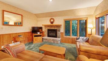 A cozy living room with a stone fireplace, wooden furniture, and large windows offering a view of trees.