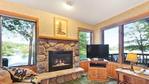 Una sala de estar acogedora con chimenea de piedra, televisor de pantalla plana y ventanas amplias que dan a un lago y árboles.