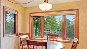 A dining area with a round table and wooden chairs, large windows with a view of a lake and trees, and a hanging light fixture. - Chanticleer Inn Villa 4 (Eagle River)