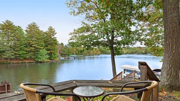 A wooden deck with wicker chairs and a glass table overlooking a lake, surrounded by trees and docked boats. - Chanticleer Inn Villa 4 (Eagle River)