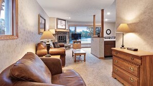 A living room with a brown leather sofa, a wooden coffee table, and a wooden dresser with drawers. There is a fireplace with a TV on top, and a large window with a view of trees and a lake. - Chanticleer Inn Villa 5 (Eagle River)