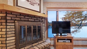 A fireplace with a stone surround, a television on a wooden stand, and a window with a view of a lake and trees. - Chanticleer Inn Villa 5 (Eagle River)