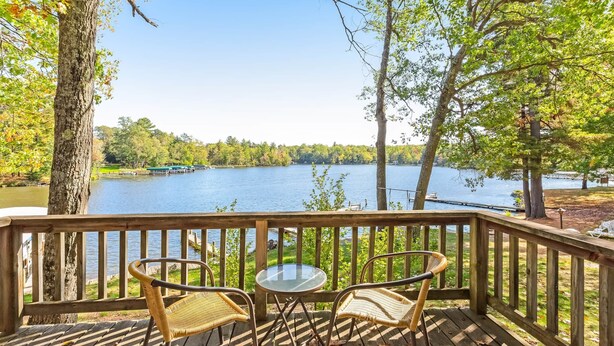 Une terrasse en bois, avec des chaises en osier et une table en verre, donnant sur un lac et entourée d’arbres.