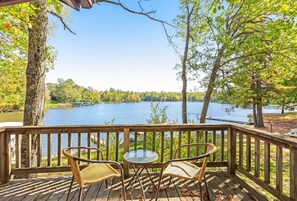 Une terrasse en bois, avec des chaises en osier et une table en verre, donnant sur un lac et entourée d’arbres.