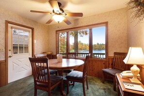 A dining area with a round wooden table and chairs, a ceiling fan, and large windows offering a view of trees and a body of water. - Chanticleer Inn Villa 7 (Eagle River)