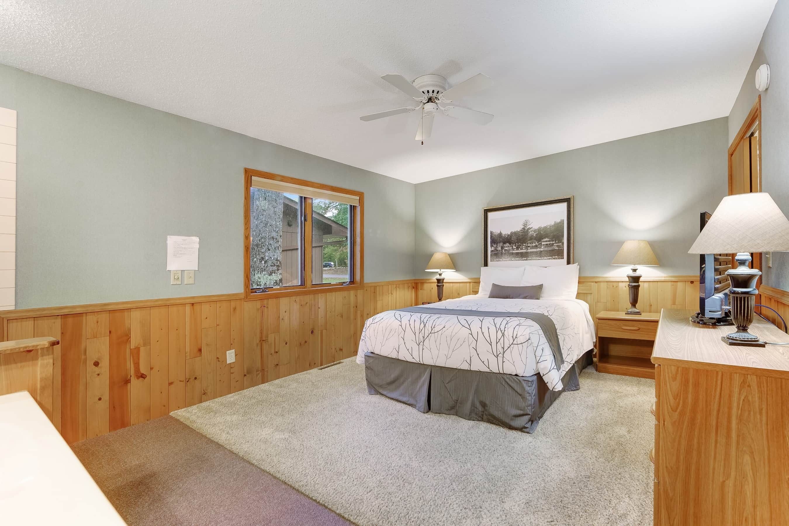 A bedroom with a bed, two bedside tables with lamps, a ceiling fan, a window, and a framed picture on the wall.