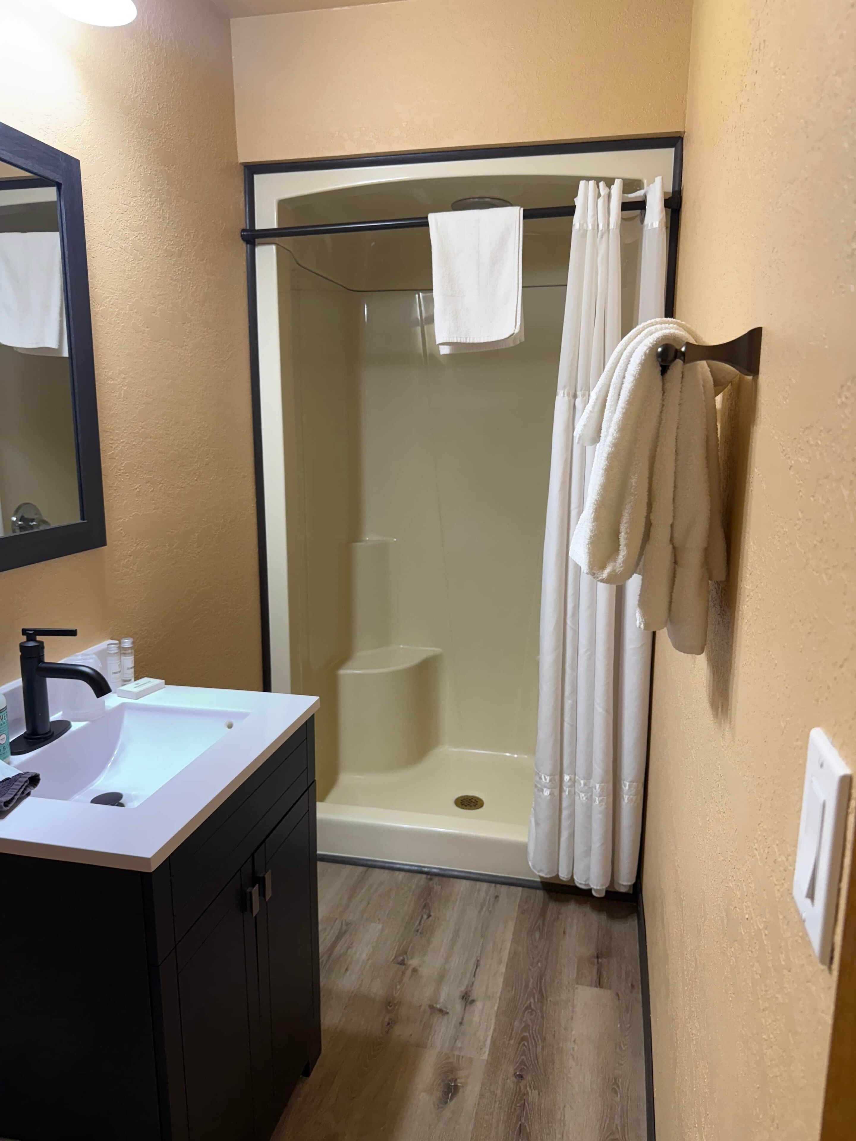 A bathroom with a white sink, black faucet, and a mirror. There is a glass shower enclosure with a white towel hanging on the door.