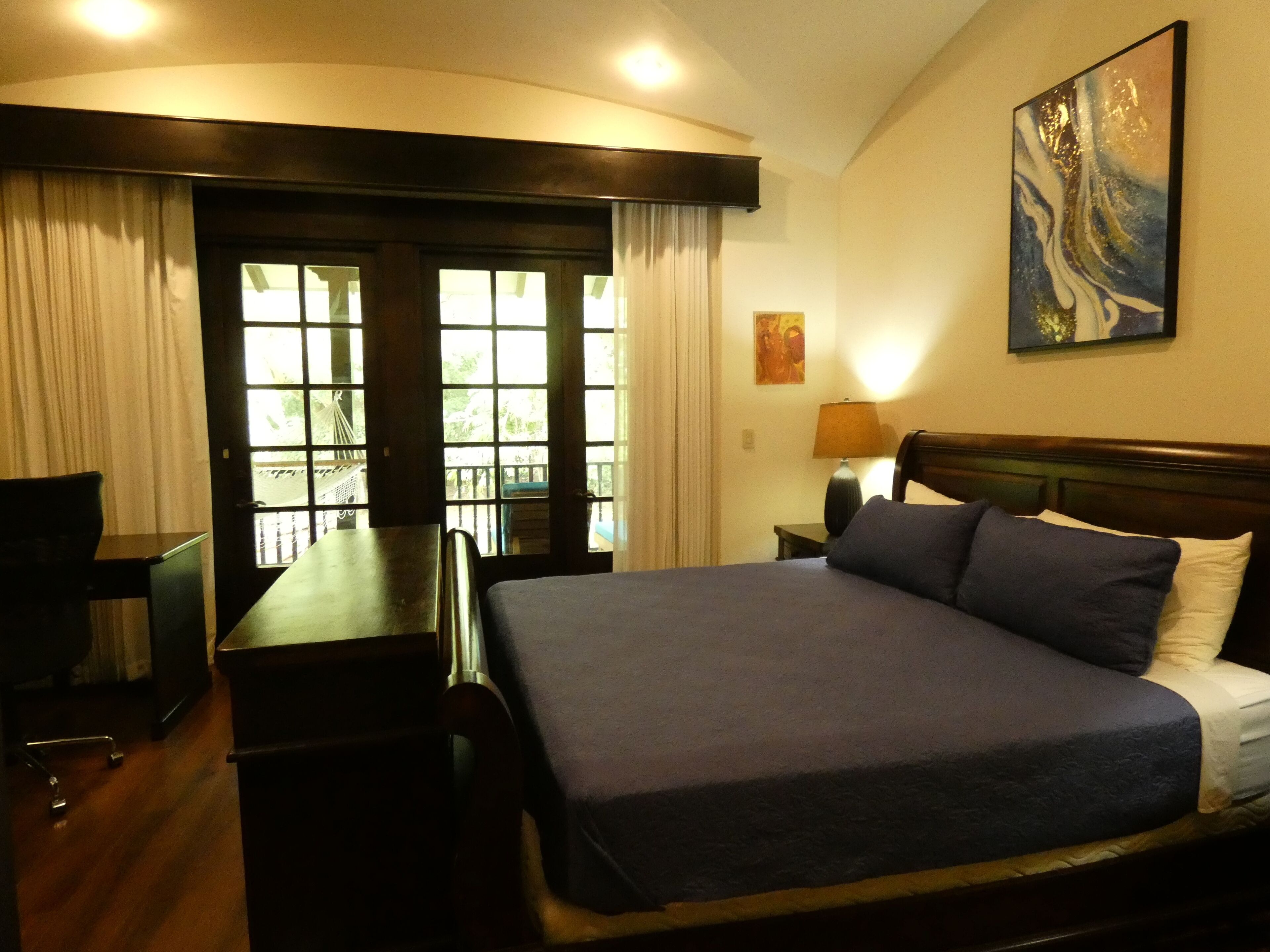 A hotel room with a large bed, a desk, and a TV. There is a window with curtains and a painting on the wall.