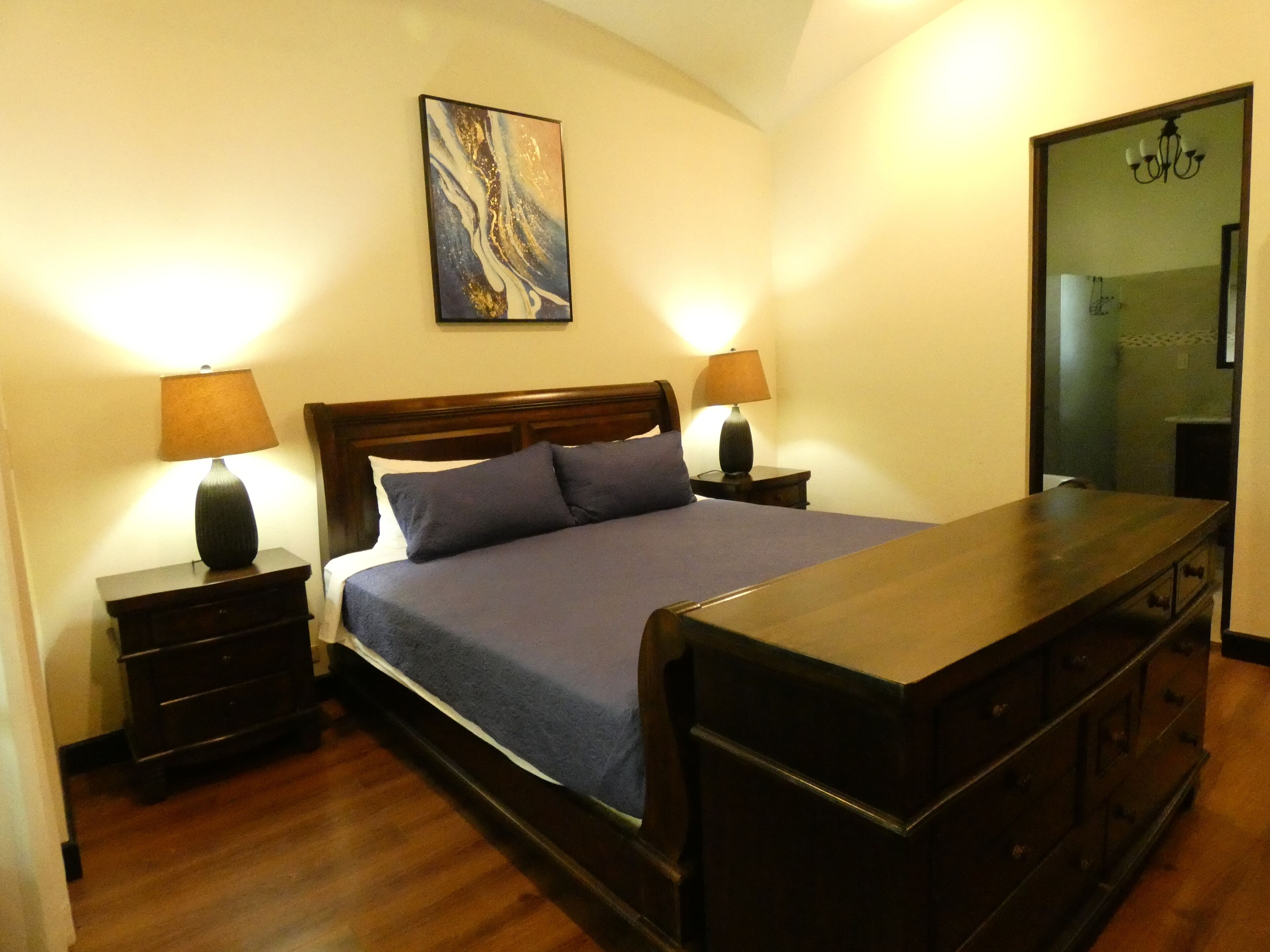 A bedroom with a wooden bed, two bedside tables with lamps, a dresser, and a painting on the wall.
