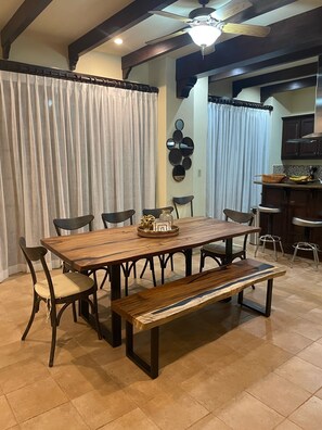 A dining area with a wooden table, six chairs, and a bench. There is a ceiling fan above and a kitchen area in the background.