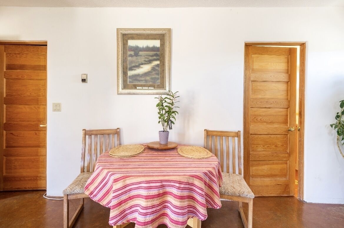 A dining area with a table covered by a striped tablecloth, two chairs, a potted plant, and a framed picture on the wall.