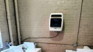Une petite buanderie équipée de deux machines à laver et d’un sèche-linge, avec un mur en briques et des tuyaux apparents.