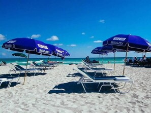 A beach with rows of sun loungers under blue umbrellas, a clear sky, and the ocean in the background.