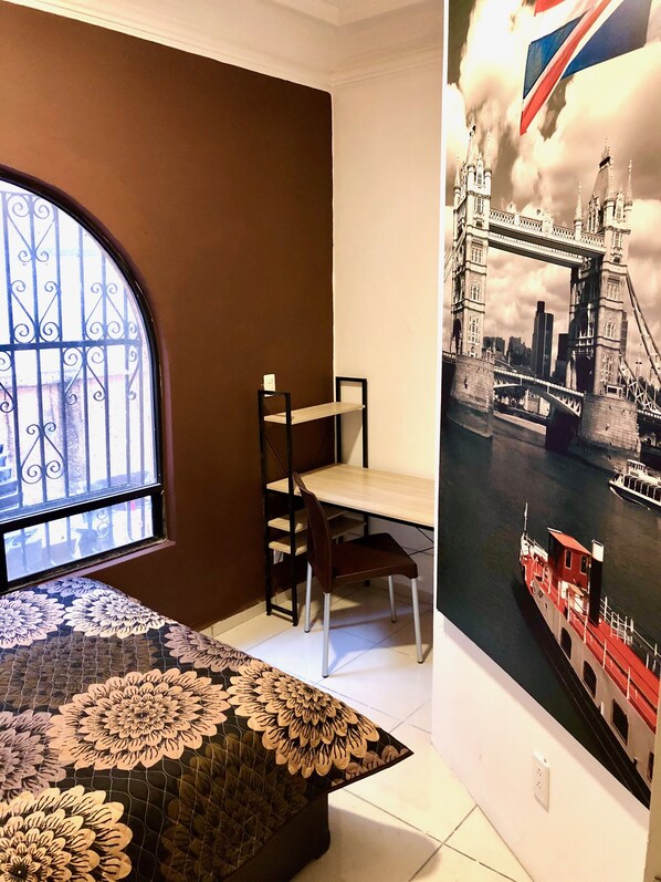 A room with a large window, a bed with a floral bedspread, a desk with a chair, and a wall mural of a bridge. - ROOMIES HOSTEL CENTRO MEDICO (Mexico City)