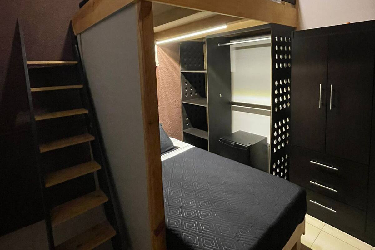 Ein Stockbett mit einer Leiter, ein Schreibtisch und ein Kleiderschrank in einem Raum mit schrägem Dach und einer Lichtleiste entlang des Randes.