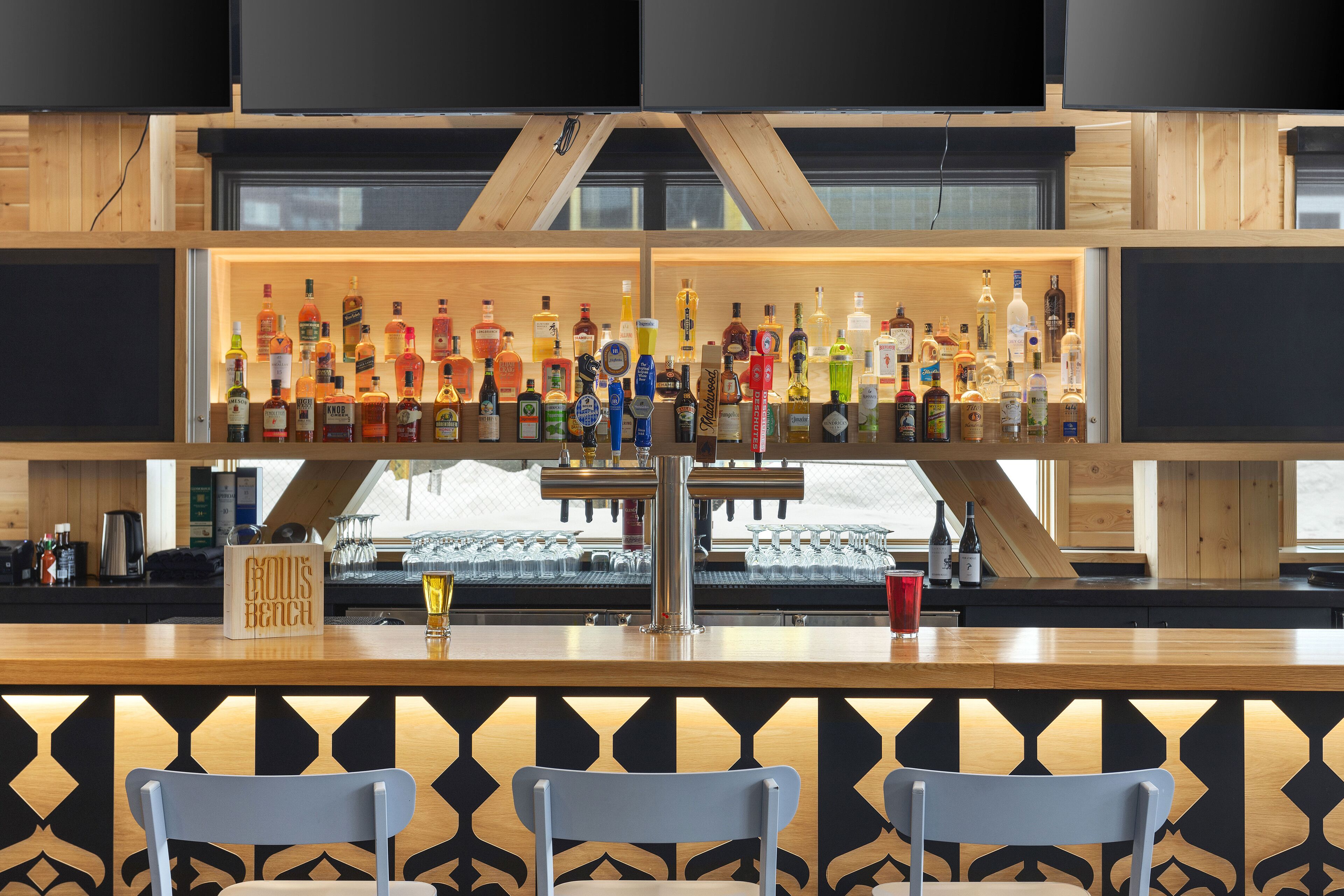 A bar area with a wooden counter, multiple liquor bottles, glasses, and a bar stool.