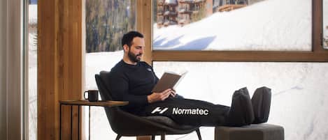 A man sitting on a chair reading a book in a room with large windows overlooking a snowy mountain landscape.