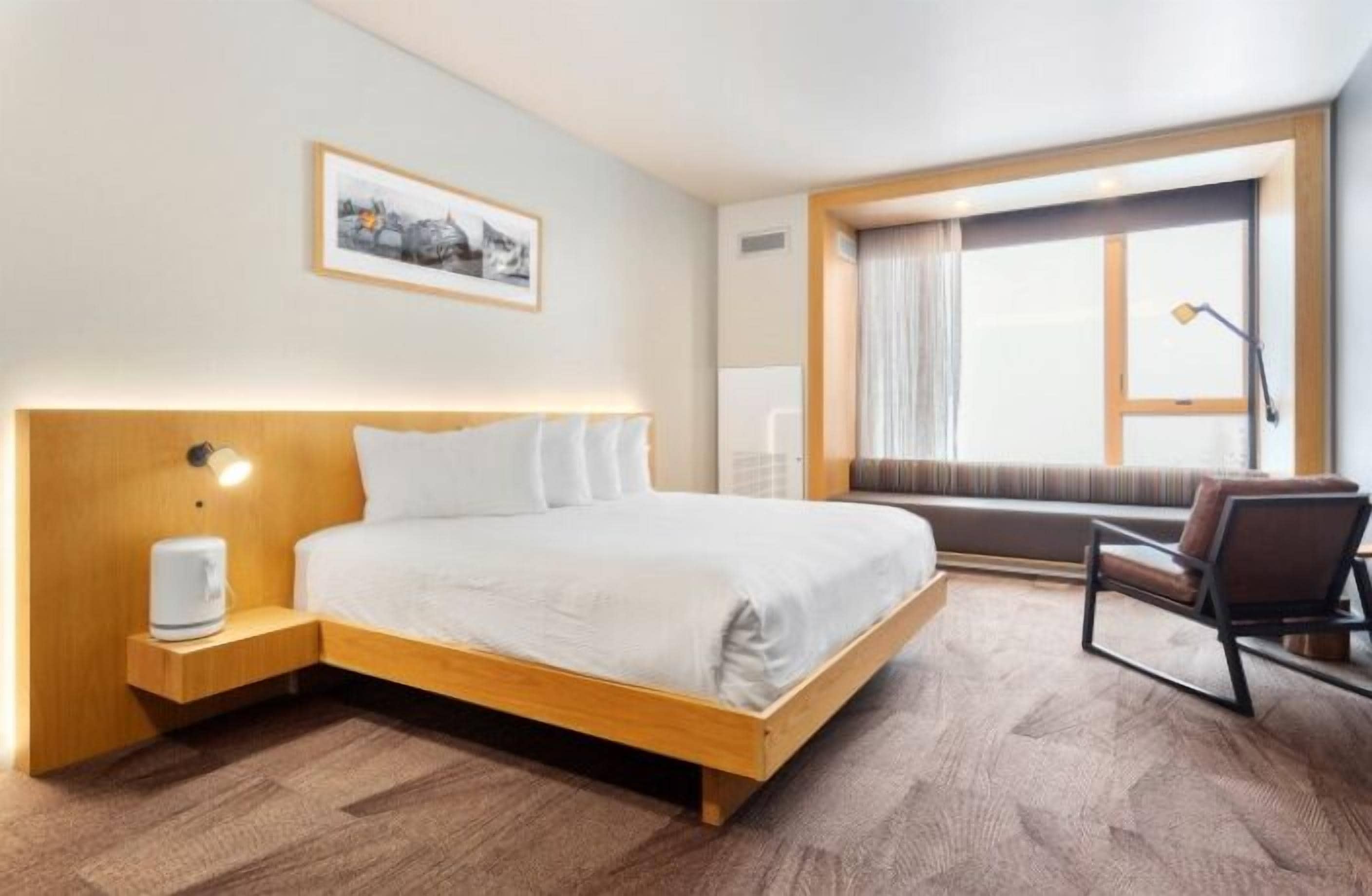 A hotel room with a wooden bed, a bedside table with a lamp, a window with curtains, and a chair.