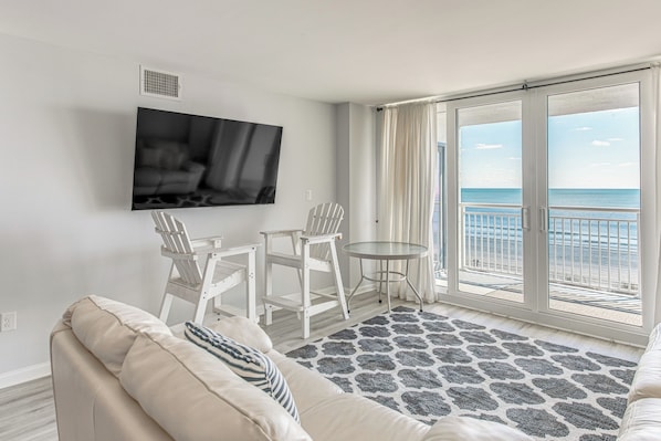 Uma sala de estar moderna com uma televisão grande, móveis brancos, uma varanda com vista para o mar e um tapete estampado.