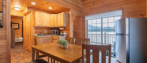 A wooden cabin interior with a kitchen, dining area, and a view through a large window.