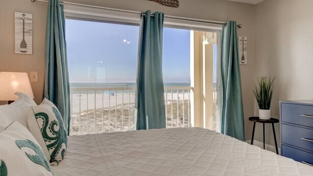 A bedroom with a bed, a nightstand, a dresser, and a view of the beach through large windows.